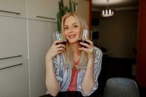 Blond woman  tases  two glasses of red wine  on her own kitchen. photo