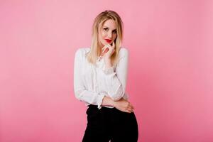 Indoor studio portrait of elegant blonde woman in white blouse and black trousers.  Posing on pink background. Red lips. photo