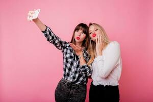 Two pretty girls making self portrait by mobile phone.  Smiling, having fun, Pink background. Studio shot. photo