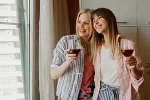 Couple of happy carefree women looking at window and holding glass of wine. Cozy home atmosphere. photo