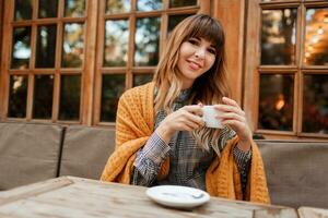 Lovely  woman have a coffee break in cozy  cafe with wood interior, talking by mobile phone. Holding cup of hot cappuccino. Winter season. Wearing elegant dress and yellow plaid. photo