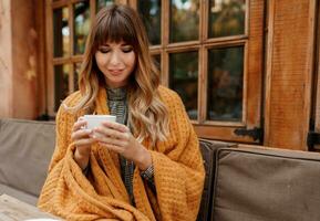 Warm cozy portrait of happy dreamy woman with wavy hairs, covered by  yellow  plaid and holding cup of hot cappuccino.  White female chilling on terrace. photo