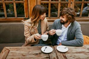 romántico mujer con largo ondulado pelos abrazando su marido con barba. elegante Pareja sentado en café con caliente capuchino. foto