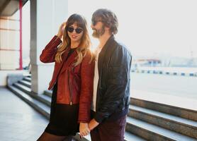 Close up fashion romantic portrait of young pretty couple kissing and hugging   on the street at evening  sunlight. Wearing leather jackets and sunglasses. Urban background. photo