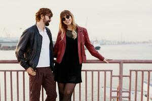 Outdoor fashion image of  stylish couple in casual outfit, leather jacket and sunglasses standing on the bridge.   Handsome man with beard with his girlfriend spending romantic time together. photo