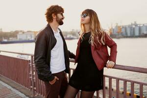 al aire libre Moda imagen de elegante Pareja en casual atuendo, cuero chaqueta y Gafas de sol en pie en el puente. hermoso hombre con barba con su Novia gasto romántico hora juntos. foto