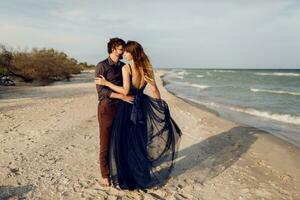 Full height image of romantic  couple embrace on the evening beach near  ocean. Stunning woman in blue long dress hugging her boyfriend with tenderness. Honeymoon. photo