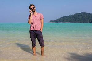 Happy  traveling man with beard  talking by mobile phone and staying  in blue sea water on his tropical  holidays. Holding  phone in hands. Full length. photo