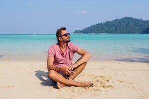 Happy attractive man  man in stylish sunglasses with beard using mobile phone , texting message. amazing blue sea background. Listening  favorite music. photo