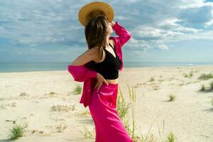 al aire libre verano imagen de elegante morena de viaje mujer posando en el playa. vistiendo elegante rosado atuendo. Paja sombrero. foto