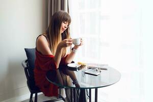 retrato de joven bonito mujer Bebiendo café , comiendo desayuno a hogar en mañana, todavía vida foto. tecnología y comodidad concepto. mirando en móvil teléfono. foto