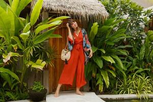 Outdoor summer fashion photo of gorgeous woman in boho   outfit posing in tropical luxury resort.  Full lenght. Tropical plants.