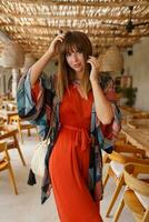 Attractive woman in orange trendy  dress  posinng in tropical cafe.  Bright outfit. Travel and vacation  fashion concept. photo