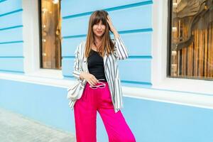 Styish woman in  striped jacket and pink pants posing on the street. Trendy summer look. Blue wall background. photo