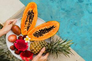 Woman holding plate of tasty  tropical exotic fruits  on the edge of pool, breakfast at luxury hotel. Slices of papaya, dragon fruit, passion fruit. Vegan and healthy food. photo