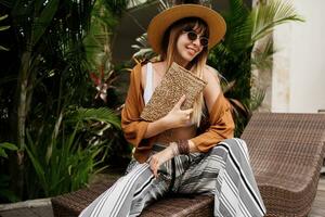 Stylish woman in summer clothes relaxing in hotel and enjoying Trendy sunglasses, straw hat and handbag, bohemian bracelets and accessorizes. photo