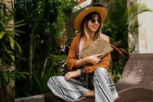 Stylish woman in summer clothes relaxing in hotel and enjoying Trendy sunglasses, straw hat and handbag, bohemian bracelets and accessorizes. photo