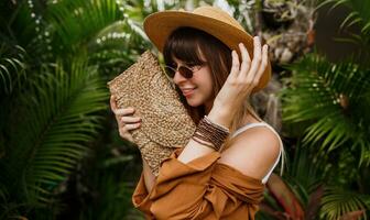 cerca arriba verano de moda retrato de morena mujer en Paja sombrero posando en tropical palma hojas antecedentes en bali vistiendo elegante bohemio accesorios. foto