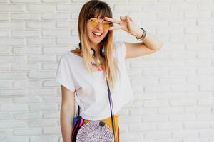 sonriente mujer posando terminado blanco ladrillo pared con auriculares y demostración señales. verano estilo. positivo humor. amarillo Gafas de sol. foto