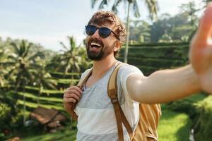 Happy stylish caucasian man with backpack travel in rice plantation and making self portrait  for memories. Concept of active lifestyle and travelling in vacation. photo