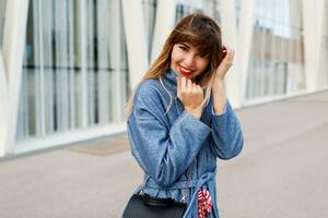 Happy brunette woman in blue coat and red dress walking along modern street , windy weather, cold season . Red lips, trendy bag. photo