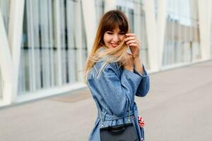 Happy brunette woman in blue coat and red dress walking along modern street , windy weather, cold season . Red lips, trendy bag. photo