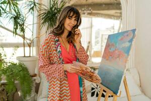 Beautiful artist woman in bohemian outfit posing with brush and palette in her art studio. photo