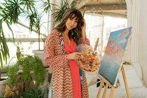 Beautiful artist woman in bohemian outfit posing with brush and palette in her art studio. photo