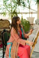 Female artist painting on canvas in her art studio. Wearing elegant boho outfit. photo