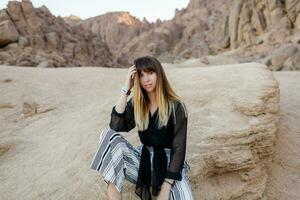 Brunette  woman  posing  in the Egyptian  desert sand dunes. Cliffs and mountains on background. Wearing black blouse and striped pants. photo