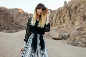 Pretty smiling fashionable girl posing  in the Egyptian  desert sand dunes. Cliffs and mountains on background. Wearing black blouse and striped pants. photo