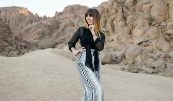 Pretty smiling fashionable girl posing  in the Egyptian  desert sand dunes. Cliffs and mountains on background. Wearing black blouse and striped pants. photo