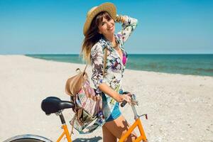 al aire libre verano imagen de bonito contento mujer montando bicicleta en increíble playa . vistiendo brillante camisa, Paja sombrero, boho mochila. foto