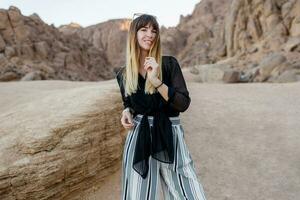 Elegant brunette woman  posing  in the Egyptian  desert sand dunes. Cliffs and mountains on background. Wearing black blouse and striped pants. photo