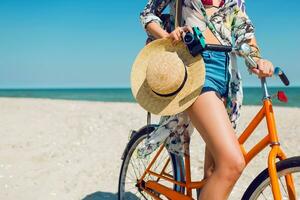 joven contento mujer en verano ropa de playa y Paja sombrero con mochila posando con Clásico bicicleta en soleado tropical playa. participación cámara. bonito turista disfrutando vacaciones. foto