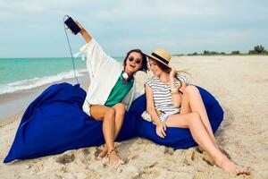 Two girls, best  friends siting on  beach pillows , having fun, enjoying  time together. Wearing stylish boho  outfit , straw hat . photo