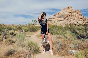 Young slim  stylish traveling woman with back pack posing near rocky mountains  in Joshua Tree National Park, California, USA. photo