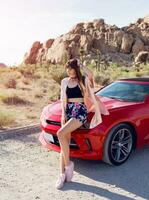 Fashionable image of pretty seductive girl sitting on  red modern sport convertible car. Joshua tree national park background. photo