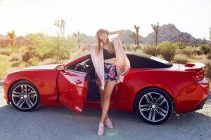 Fashionable image of pretty seductive girl sitting on  red modern sport convertible car. Joshua tree national park background. photo