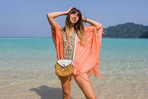 al aire libre verano retrato de elegante bronceado pensativo mujer en de moda boho playa vestir con bordado y borla. Paja bolsa. foto