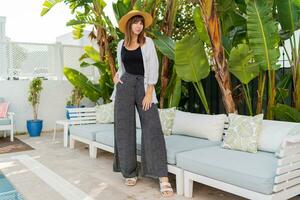 Elegant woman in straw hat posing  in cozy tropical resort near pool. Tropical leaves on background. Stylish white furniture. Full lenght. photo