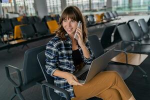 Airport Young female passenger  with smart phone and laptop sitting in terminal hall while waiting for her flight photo