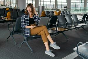 Young female passenger with  laptop sitting in terminal hall while waiting for her flight photo