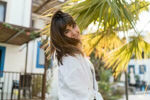 alegre morena mujer disfrutando verano día. vistiendo blanco blusa. palma arboles en antecedentes. foto