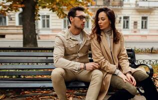 Brunette model with stylish guy in beige coat sitting on bench. photo