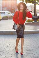 seductive brunette woman in autumn casual outfit walking in sunny city . Red knitted pullover, black trendy hat, leather skirt. photo