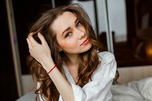 Close up lifestyle  morning portrait of pretty young woman sitting on bed . Wearing white    shirt and have wavy long hair.  Positive happy mood. photo