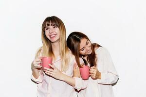 Two cheerful  white   women  in pink pajamas  with cup of tea posing on white background. Flash portrait. photo