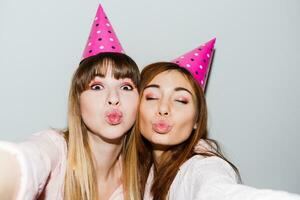 Self portrait of two smiling women in pink  paper birthday hats  on white background.  Friends wearing pink pajamas. photo