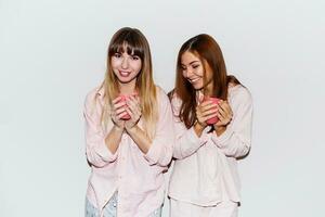 Two cheerful  white   women  in pink pajamas  with cup of tea posing on white background. Flash portrait. photo